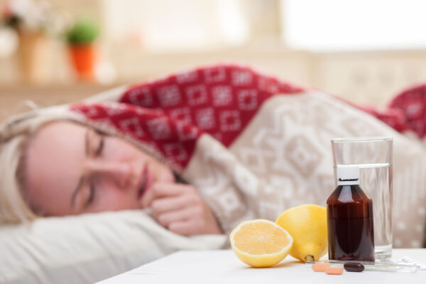 Ill young woman is sleeping in bed. She has a fever. Focus on pills, lemon and a glass of water on the table